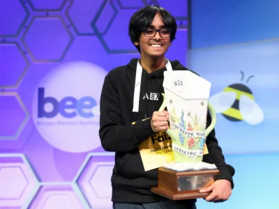 Young Scripps National Spelling Bee champion smiling on stage while holding the championship trophy in front of the official Bee backdrop during the 2026 finals competition