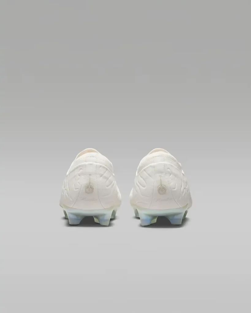 Rear view of a pair of premium football boots shown in an all-white finish with sculpted embossed detailing wrapping around the heel. The back panel features subtle tonal branding and signature-style script above the heel counter, while the upper extends into a softly padded low-cut collar. Beneath, a translucent iridescent soleplate with pearl-like blue reflections and angular heel studs adds contrast to the otherwise monochromatic design. Displayed symmetrically against a clean grey studio backdrop, the design emphasizes refined craftsmanship and performance-driven construction