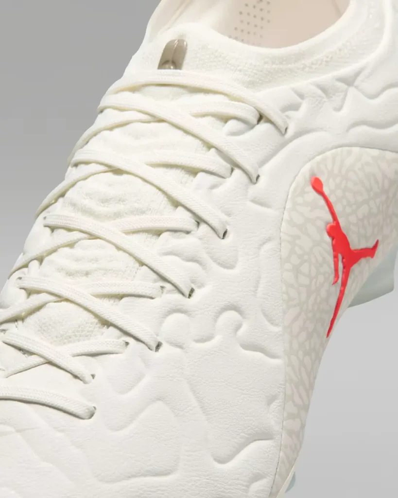 Close-up detail of a premium football boot in an all-white finish highlighting the textured upper, tonal lacing system, and sculpted organic embossing across the forefoot and midfoot. A cracked elephant-inspired panel wraps the lateral side and is contrasted by a glossy vivid red Jumpman logo. Fine suede-like textures and layered material transitions emphasize the crafted, luxury-inspired aesthetic, while hints of the iridescent soleplate appear beneath against a clean grey studio background