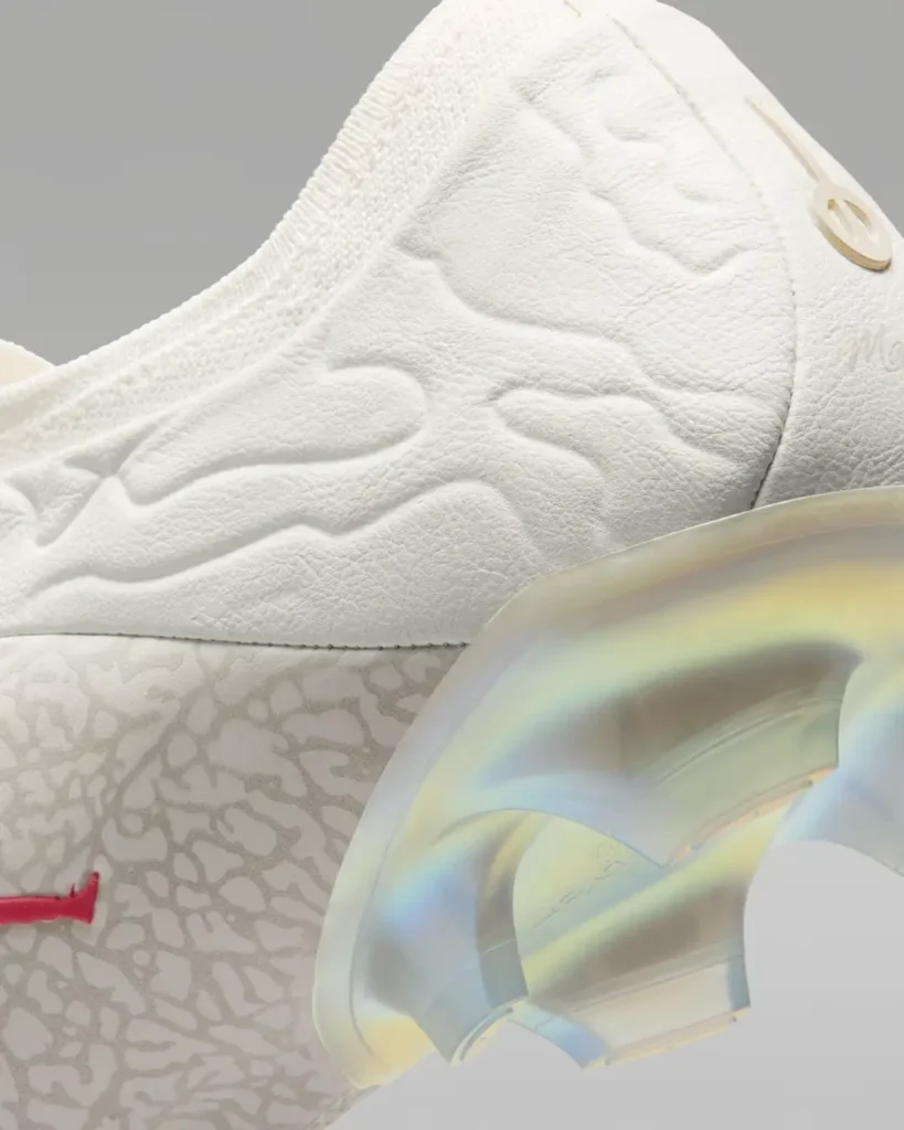 Macro close-up of the heel and rear-quarter area of a premium football boot in an all-white finish, showcasing layered material textures and embossed organic contours across the leather upper. A tonal cracked elephant-inspired overlay wraps the lower heel, accented by a partial glimpse of a vivid red Jumpman logo. The iridescent translucent heel plate catches blue, gold, and pearl reflections, emphasizing the contrast between soft tactile materials and the sculpted performance sole construction against a neutral grey studio backdrop