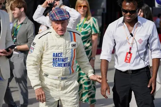 Jackie Stewart walks through a paddock-style setting wearing a cream racing suit paired with a colorful plaid flat cap, blending heritage style with motorsport authenticity. The suit features bold blue text reading “Race Against Dementia” across the chest, along with technical patches such as OMP branding and vertical stripe detailing in red and blue along one side. His look is complemented by casual confidence and a slightly animated stride, as he gestures mid-step. Beside him, another man in a white button-down shirt, dark trousers, and sunglasses wears a paddock credential lanyard, suggesting an official or team role. In the background, a small crowd moves through what appears to be a race-day environment, reinforcing the live-event atmosphere. The overall scene captures a crossover between racing heritage, advocacy, and contemporary F1 culture.