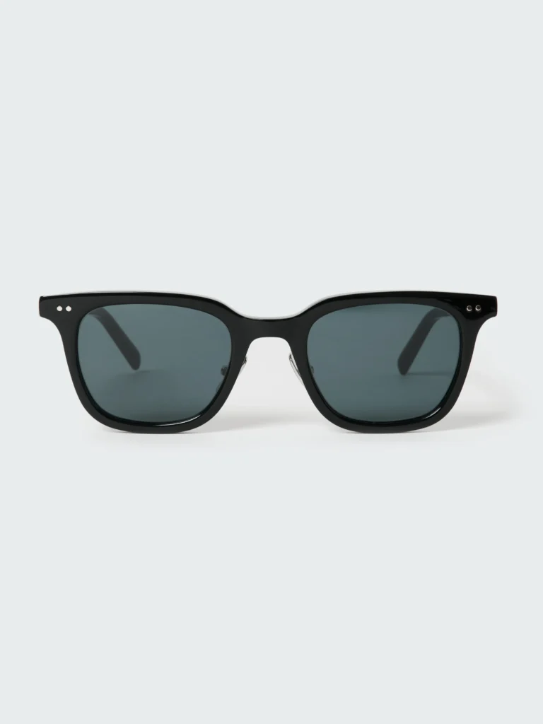 Minimal black square-frame sunglasses with thick bio-circular acetate construction, dark tinted lenses, and small silver rivet details at the corners, photographed against a soft light gray background