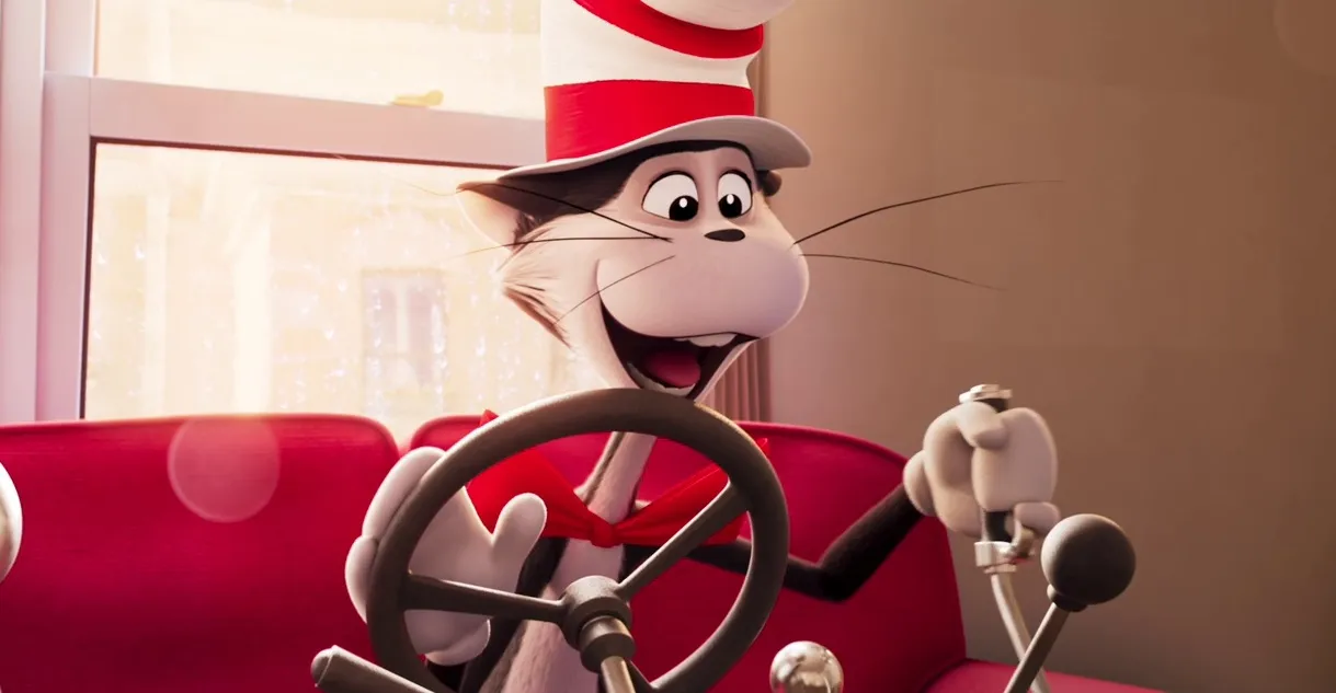 Animated still from The Cat in the Hat (2026) showing the iconic Dr. Seuss character enthusiastically steering a whimsical vehicle inside a brightly lit room. Wearing his signature red-and-white striped hat and red bow tie, the Cat smiles broadly while seated against a vivid red sofa, highlighting the film’s colorful, play, animation style