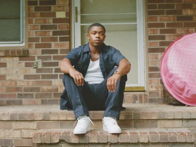 A portrait of Trap Dickey seated on the brick steps of a modest Southern-style home, wearing a dark navy workwear-inspired set layered over a white tank top with crisp white sneakers. The composition carries a quiet, reflective atmosphere, with muted film-like tones and natural daylight softening the scene. A faded pink plastic kiddie pool at the side subtly contrasts the earthy brick textures, reinforcing the image’s grounded neighborhood realism