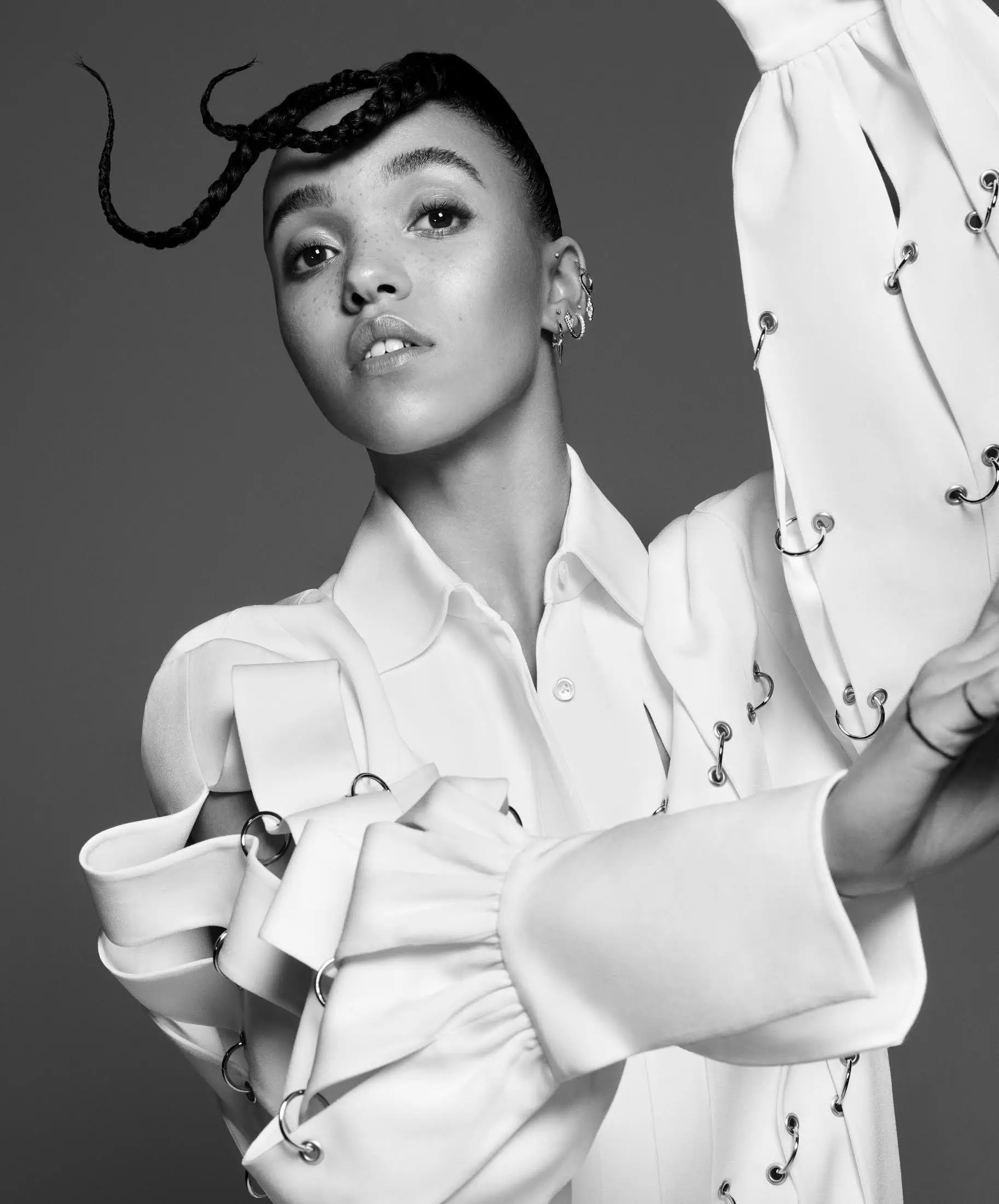 Black-and-white fashion portrait of a model posing in an avant-garde white blouse detailed with metallic ring fastenings and exaggerated sculptural sleeves, complemented by braided hair styling, subtle freckles, layered ear piercings, and a poised editorial expression against a minimalist studio backdrop