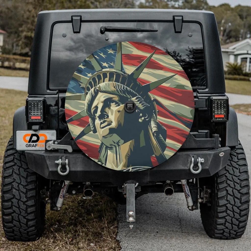 Rear view of a customized black Jeep Wrangler parked roadside featuring a full spare tire cover printed with a stylized illustration of the Statue of Liberty layered over a flowing American flag motif. The artwork uses muted patriotic tones of red, cream, blue, and olive in a graphic poster-like treatment. Rugged off-road tires, exposed tow hooks, and aftermarket lighting reinforce the Jeep’s utilitarian character while the illustrated spare cover becomes the view centerpiece