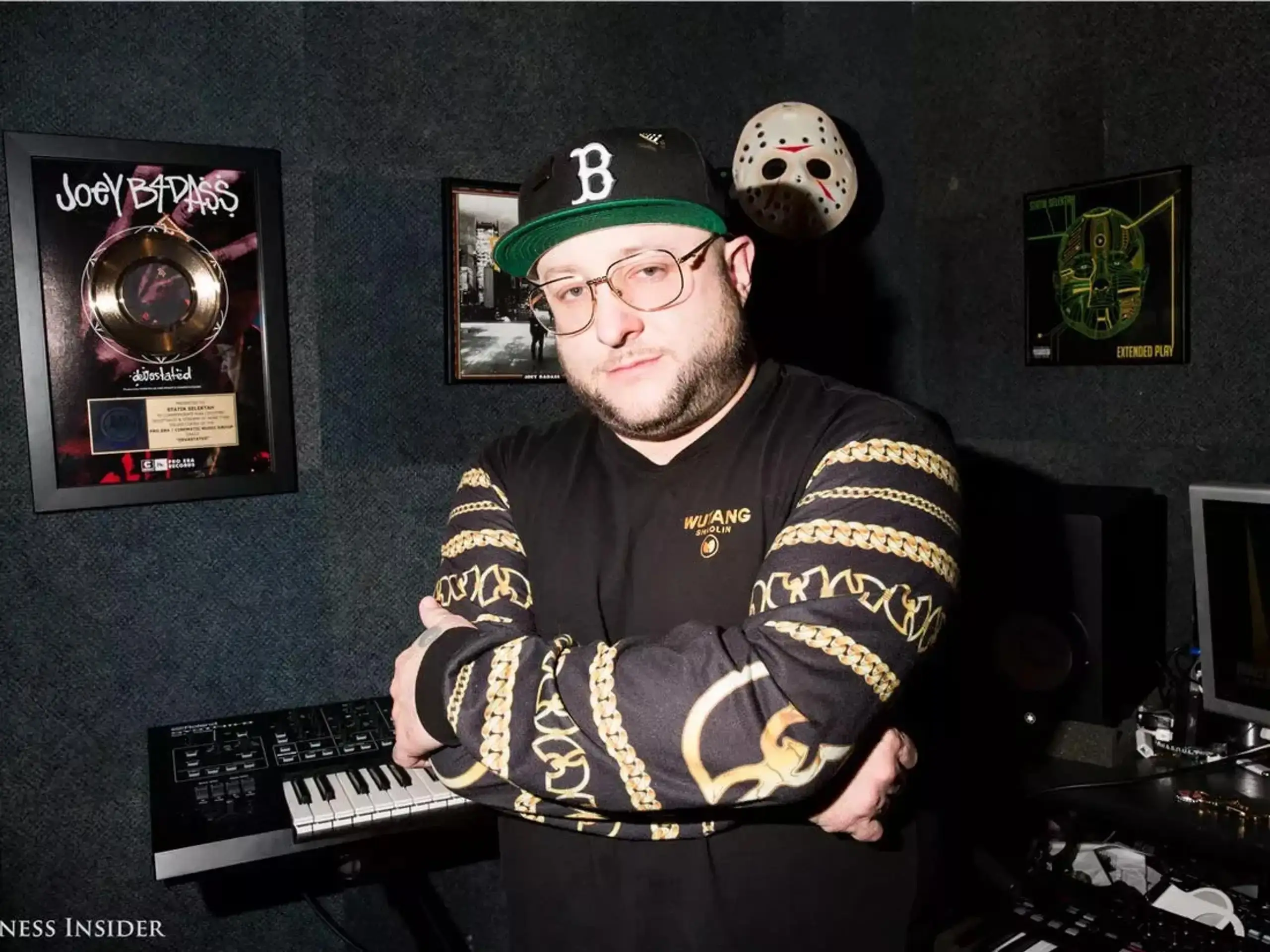 Man in a dark music studio wearing a black “B” cap and gold-patterned Wu-Tang shirt, standing with arms crossed in front of a keyboard, framed Joey Bada$$ plaque, wall-mounted hockey mask, and studio equipment