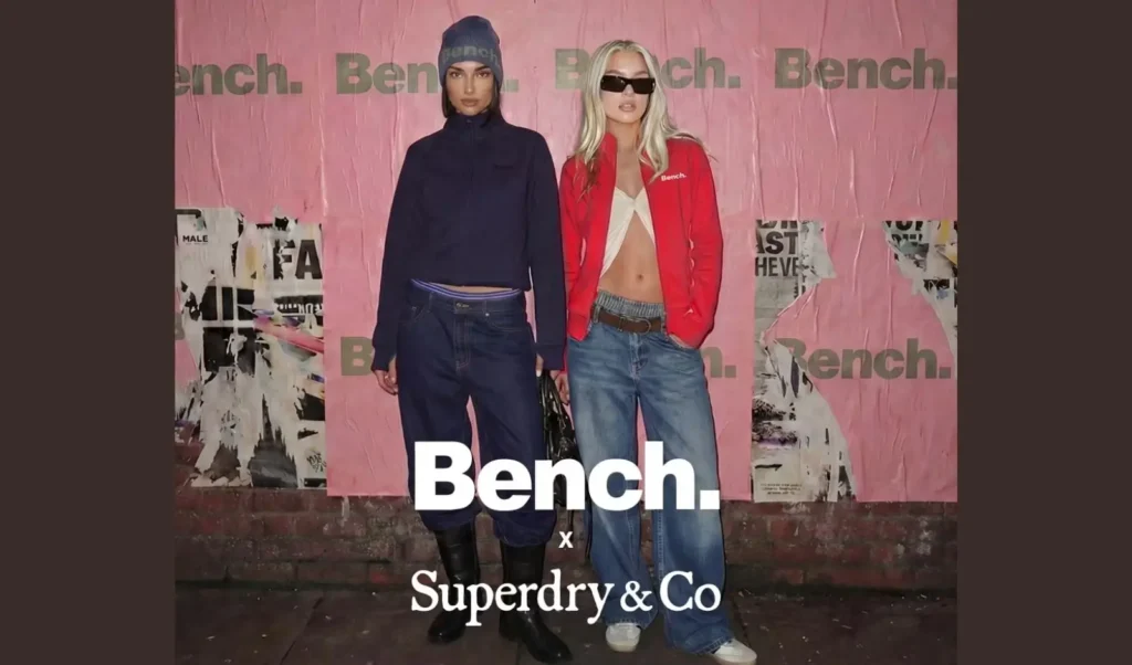 image for the Bench x Superdry & Co. relaunch campaign featuring two models standing against a pink Bench-branded backdrop with distressed poster details. One model wears a navy cropped zip jacket with matching beanie and dark denim, while the other wears a red Bench zip jacket with loose blue jeans and black sunglasses. Large white “Bench.” and “Superdry & Co” logos overlay the image, emphasizing the brand’s Y2K-inspired streetwear revival