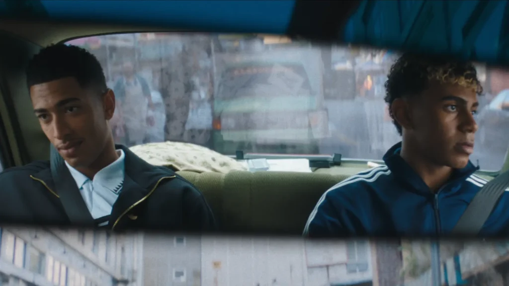Two young men sit in the back seat of a car in a cinematic wide shot framed through the vehicle’s rearview mirror. One wears a dark adidas track jacket layered over a crisp white collared knit, while the other sports a royal blue adidas zip-up with classic white three-stripe detailing and a seatbelt crossing his chest. Soft daylight filters through fogged windows, revealing blurred urban streets outside and creating a reflective, intimate atmosphere. The muted tones, casual posture, and understated sportswear styling blend contemporary football culture with nostalgic city realism and elevated adidas aesthetics