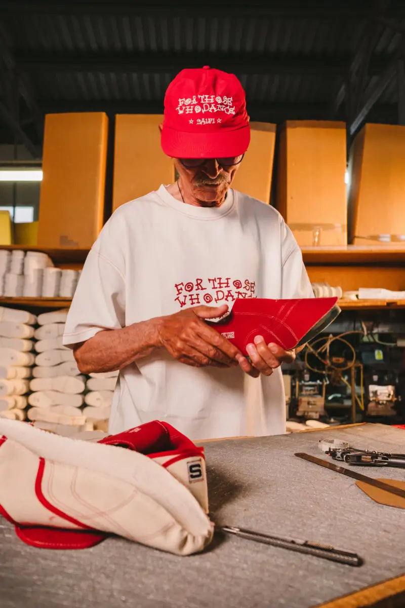 Elder artisan in a workshop examining a red Victoria x SALPI vulcanised boot while wearing matching “For Those Who Dare” apparel, surrounded by shoe materials and production tools in a warm industrial setting