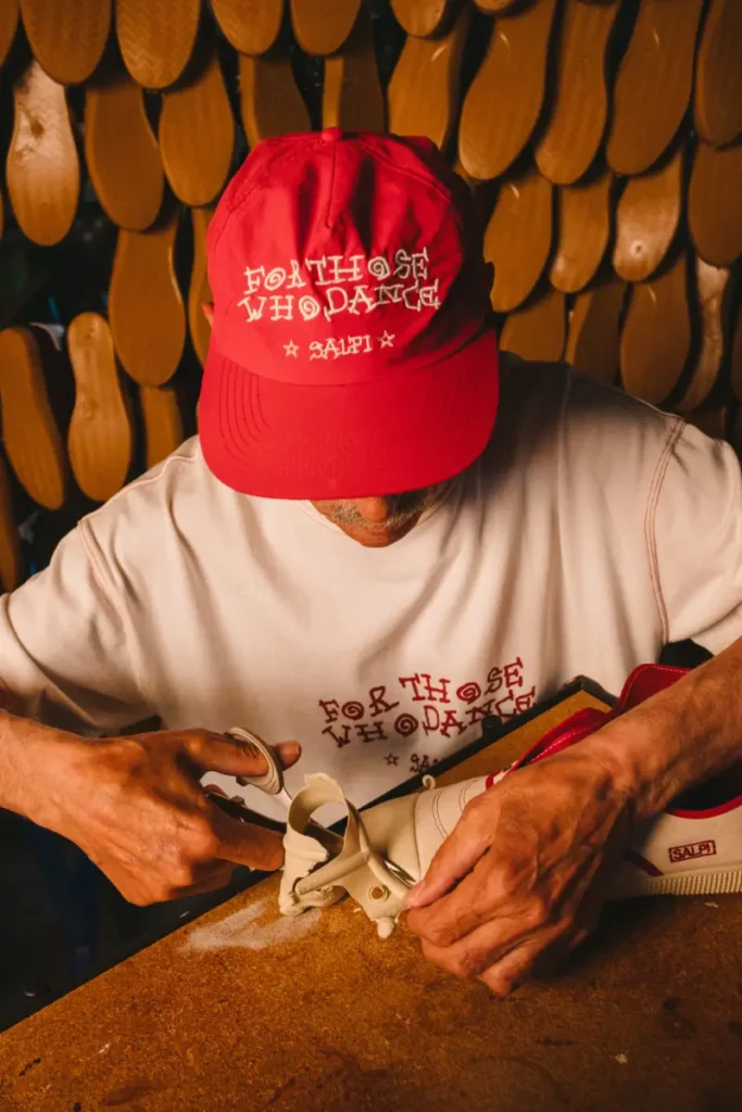 Artisan wearing SALPI collaborative apparel carefully cutting and shaping a Victoria vulcanised shoe by hand, surrounded by stacked rubber soles inside a warm-toned footwear workshop