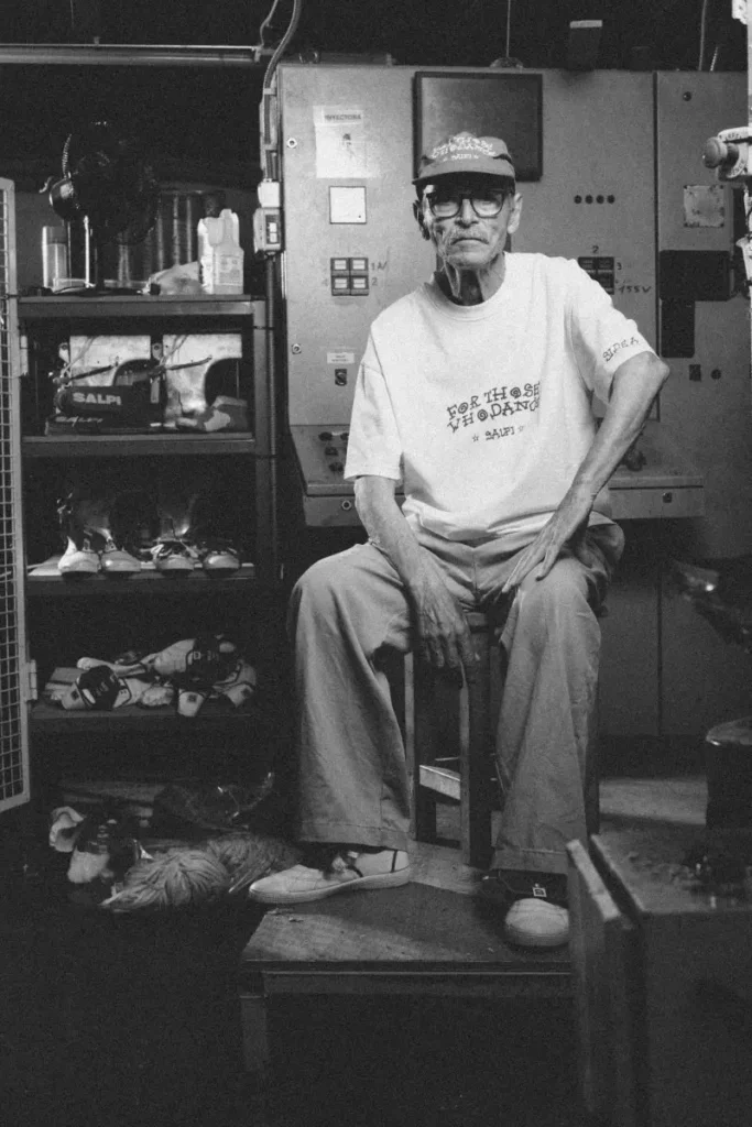 Black-and-white portrait of the SALPI founder seated inside an industrial footwear workshop wearing Victoria x SALPI apparel and shoe, surrounded by machinery, tools, and prototype shoes