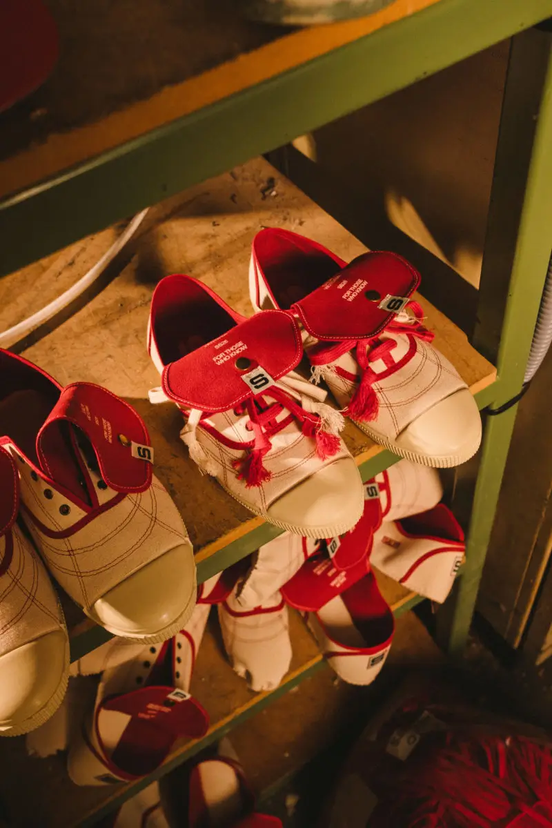 Multiple pairs of Victoria x SALPI vulcanised shoe arranged on workshop shelving, showcasing oversized red tongues, visible stitching, layered laces, and cream canvas construction under warm industrial lighting