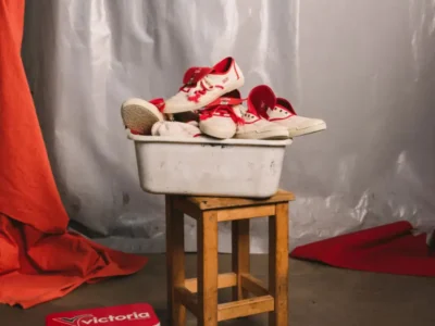 Multiple pairs of Victoria x SALPI vulcanised shoe displayed inside a worn metal basin atop a wooden stool, set against draped studio fabric with red accents and vintage Victoria packaging nearby