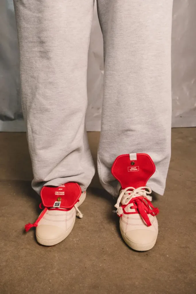 Front-facing close-up of the Victoria x SALPI vulcanised shoe featuring oversized red tongue flaps, mismatched layered laces, cream canvas uppers, and visible red stitching beneath relaxed grey sweatpants