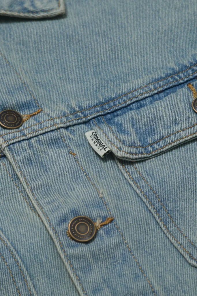Close-up detail of a light-wash denim jacket showcasing curved seam construction, contrast golden stitching, and oversized flap pockets with branded metal buttons. The faded indigo treatment and sculpted panel lines emphasize a worn-in, contemporary reinterpretation of classic utility denim
