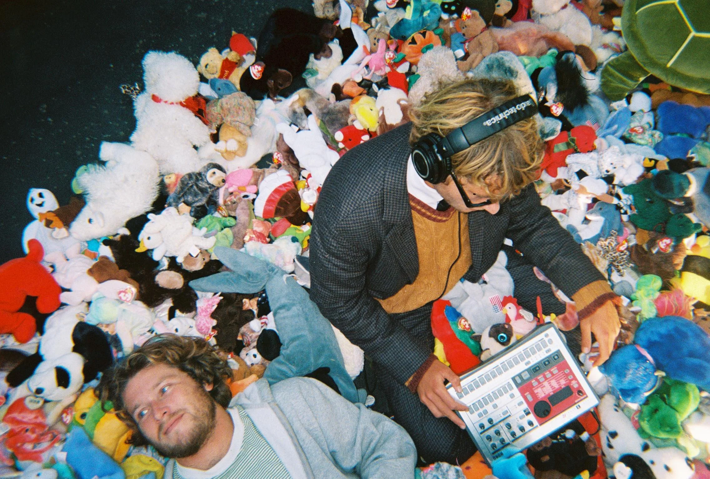 Overhead editorial-style photograph of two musicians or producers surrounded by a dense sea of colorful plush toys and stuffed animals. One person wears headphones while adjusting a compact music production device, dressed in layered knitwear and tailoring, while the other reclines among the toys. The playful composition blends lo-fi studio culture, nostalgic textures, and experimental indie aesthetics