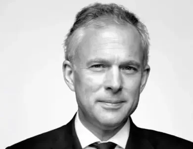 Black-and-white corporate portrait of a middle-aged man in a dark suit and tie, photographed against a plain light background with a composed expression and subtle smile