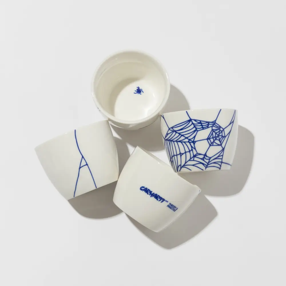 Overhead view of four white porcelain soba cups from Carhartt WIP’s “Theory and Practice” collection arranged on a light surface, featuring blue spiderweb graphics, minimalist line detailing, and subtle branding that merges Japanese-inspired ceramic tradition with contemporary streetwear design language