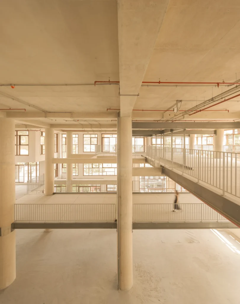 A vast open-plan architectural interior unfolds across multiple levels with exposed concrete ceilings, cylindrical support columns, and suspended walkways lined with minimalist railings. Warm natural light pours through expansive grid-framed windows, illuminating the unfinished industrial-modern space while a blurred figure crossing one of the elevated corridors emphasizes the building’s monumental scale and airy spatial composition