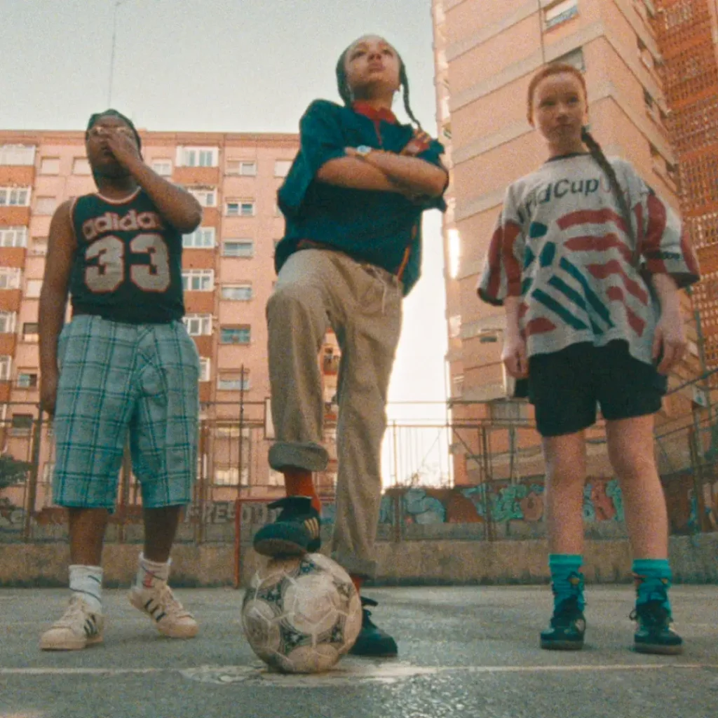 Three young people stand confidently on an outdoor urban football court framed by tall apartment buildings and graffiti-covered barriers. The central figure rests one foot on a worn football while wearing loose neutral trousers and a dark adidas top with red detailing, flanked by friends dressed in retro-inspired sportswear including a vintage adidas basketball jersey and an oversized USA World Cup graphic shirt. Black adidas sneakers, striped socks, and relaxed silhouettes reinforce the nostalgic street-football aesthetic, while warm cinematic grain and low-angle framing give the scene an energetic neighborhood-tournament atmosphere