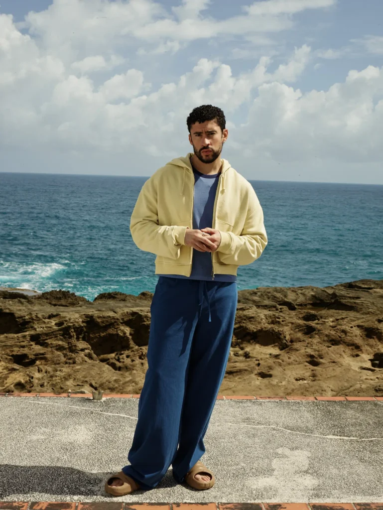 A man stands against a dramatic ocean backdrop beneath towering clouds, wearing relaxed layered separates in washed, sun-softened tones. The look combines a pale yellow cropped zip hoodie over a muted blue tee with wide-leg blue drawstring trousers and slip-on sandals, creating an effortless coastal silhouette that feels easygoing, oversized, and quietly refined