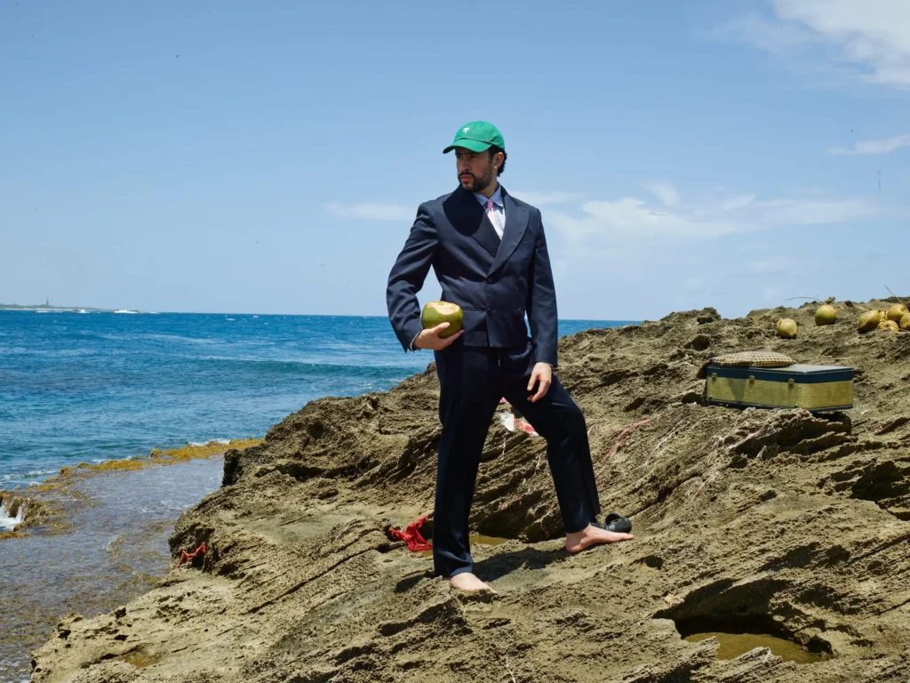 A man stands barefoot across sunlit coastal rocks overlooking the open ocean, dressed in a sharply tailored navy suit styled with a checked shirt, soft pink tie, and vivid green cap. Holding a fresh coconut while framed by scattered seaside objects and expansive blue sky, the scene blends formal dressing with relaxed island imagery in a editorial way
