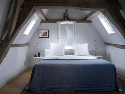 A minimalist attic bedroom is framed by exposed dark wooden beams and angled white walls, blending rustic architecture with clean contemporary design. A neatly made bed with crisp white pillows and a deep blue linen cover sits centered beneath a small window, while skylights on both sides flood the quiet space with soft natural light