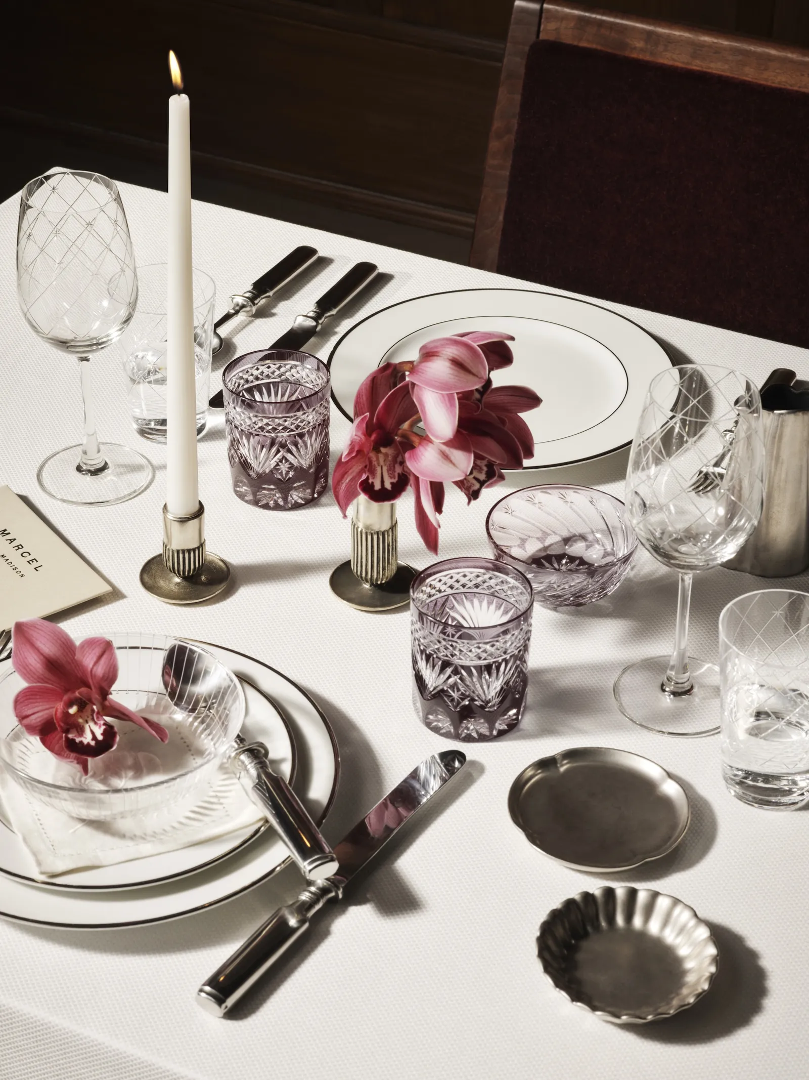 Refined Marcel table setting with crystal glassware, silver flatware, candlelight, and vibrant orchid arrangements against a warm wood backdrop
