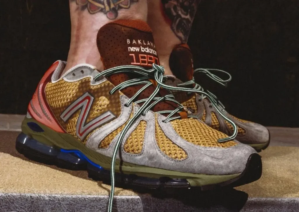 Detailed close-up of a pair of Baklava x New Balance 1890 shoe featuring olive mesh uppers, gray suede overlays, oversized mint rope laces, and perforated brown tongues with glowing orange branding. The sculpted sole unit combines translucent cushioning with earthy green and blue accents, while tattooed ankles and warm low lighting emphasize the shoe’s rugged trail-runner inspiration and collectible streetwear identity