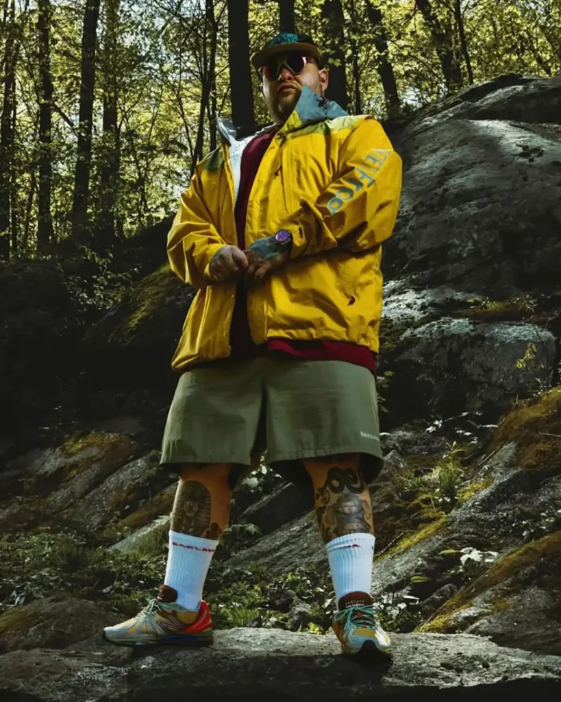 Low-angle outdoor portrait of a tattooed man standing on moss-covered rocks in a dense forest wearing a vivid yellow technical shell jacket, olive trail shorts, white crew socks, and colorful performance sneakers. A cap and dark sunglasses add to the rugged styling, while filtered sunlight through the trees creates a cinematic contrast between the bright outerwear and the shadowy woodland environment