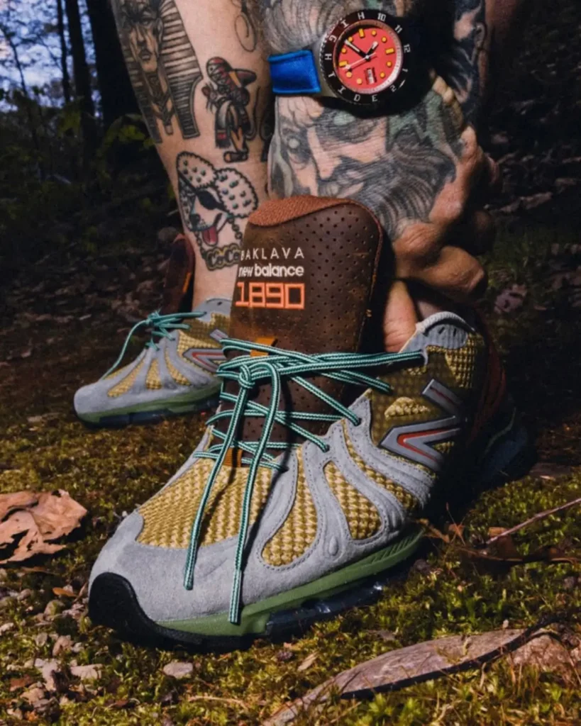 Close-up outdoor shot of heavily tattooed legs wearing textured New Balance trail sneakers in earthy olive, gray, and mint tones with oversized turquoise laces and a brown tongue branded “Baklava New Balance 1890.” A colorful wristwatch and moss-covered forest ground reinforce the rugged hiking-inspired aesthetic, while the detailed materials and layered color palette give the footwear a bold, collectible streetwear feel