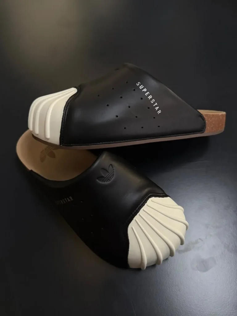 A detailed studio-style look at the adidas Originals Adimule Superstar in its black colorway highlights the silhouette’s playful hybrid construction and unmistakable Shelltoe identity. The oversized cream rubber toe cap dominates the front profile with sculptural ridges that exaggerate the classic Superstar DNA, while the smooth black upper maintains clean perforation detailing and minimalist “SUPERSTAR” branding along the lateral side. The open-back mule construction reveals a cork-inspired footbed stamped with the Trefoil logo, reinforcing the model’s relaxed, slip-on functionality. Set against a dark reflective backdrop, the composition emphasizes the balance between heritage shoe culture and contemporary comfort-driven design, positioning the Adimule Superstar as both a nostalgic icon and an easygoing summer lifestyle piece