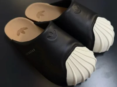 A clean product-focused view of the adidas Originals Adimule Superstar showcases the silhouette’s exaggerated Shelltoe design and relaxed mule construction in its most sculptural form. The smooth black upper is stripped back to essentials, interrupted only by subtle perforations and embossed Trefoil branding, while the oversized cream rubber shell toe becomes the unmistakable focal point with its deeply ridged, almost organic texture. The open-back structure exposes a soft cork-inspired footbed stamped with adidas Originals branding, reinforcing the shoe’s comfort-driven identity. Against the dark minimalist backdrop, the Adimule Superstar reads as a playful fusion of heritage basketball sneaker culture and modern lounge-ready footwear, balancing archival familiarity with contemporary slip-on ease