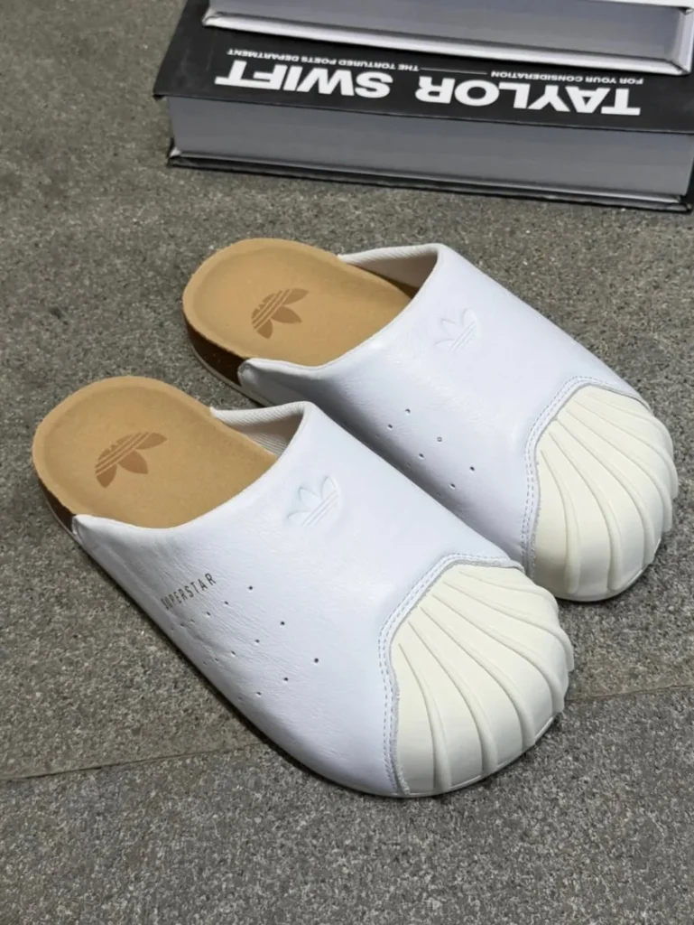 The adidas Originals Adimule Superstar appears in a clean white execution that amplifies the silhouette’s playful proportions and relaxed luxury aesthetic. The smooth white upper flows seamlessly into the exaggerated cream Shelltoe, whose sculpted ridges remain the unmistakable focal point of the design. Minimal perforation detailing and subtle Trefoil embossing preserve the classic Superstar identity while allowing the mule’s simplified form to feel contemporary and uncluttered. The cork-inspired footbed introduces a warm tonal contrast against the monochromatic upper, reinforcing the shoe’s comfort-driven, slip-on philosophy. Positioned beside stacked design books, the composition frames the Adimule Superstar less as a traditional shoe and more as a lifestyle object blending archival sportswear heritage with modern indoor-outdoor ease