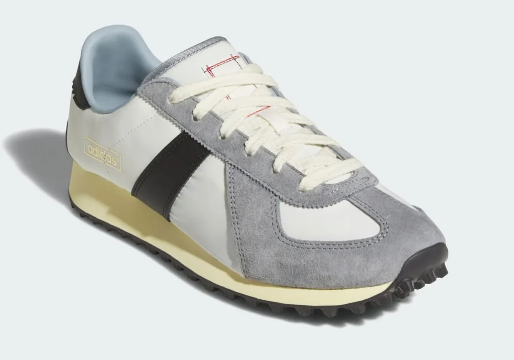 A vintage-inspired adidas running shoe presented in a clean neutral colorway featuring a white nylon upper, soft gray suede overlays, and contrasting black leather side stripes. The low-profile silhouette is accented by cream-toned laces and an aged off-white foam midsole that evoke classic marathon footwear from the late 1970s and early 1980s. Rugged black traction detailing beneath the outsole adds a utilitarian edge, while subtle gold adidas branding near the heel maintains the shoe’s understated heritage aesthetic. The combination of muted textures and archival proportions gives the sneaker a refined balance between retro sportswear nostalgia and modern everyday versatility
