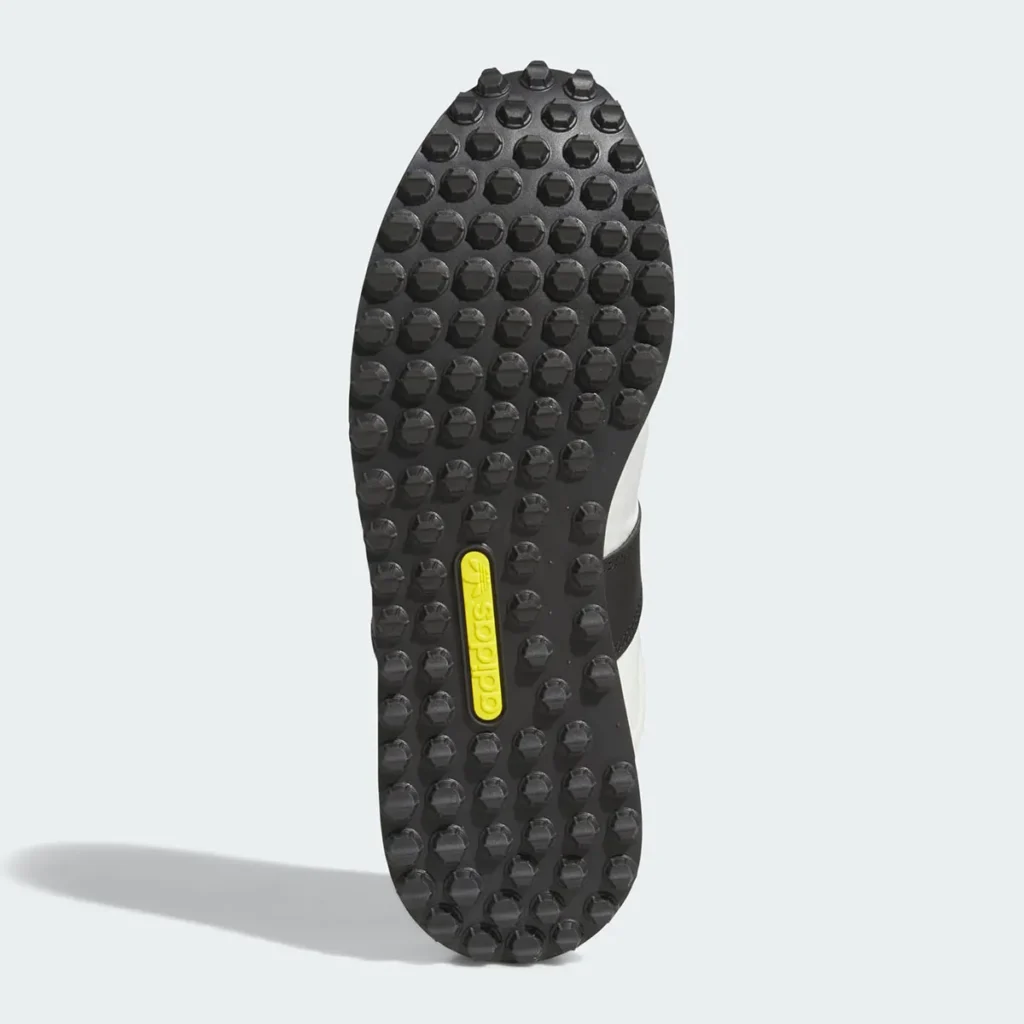 A detailed outsole view of a retro adidas running shoe highlighting its aggressive black rubber traction pattern inspired by classic archival marathon footwear. The sole features a dense arrangement of rounded lug studs designed for grip and durability, giving the sneaker a rugged vintage runner identity. At the center, a slim yellow adidas Trefoil insert introduces a subtle pop of color against the otherwise monochromatic black base, reinforcing the shoe’s heritage branding. The overall construction emphasizes functional retro engineering while complementing the silhouette’s understated lifestyle-oriented aesthetic