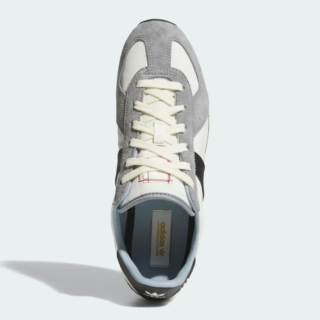 A top-down product view of a retro-inspired adidas running shoe showcasing its clean vintage construction and material layering. The shoe combines a white nylon base with soft gray suede overlays wrapping the toe and eyestay, contrasted by black leather side stripes partially visible beneath thick cream-colored laces. Subtle red stitching accents on the tongue introduce a restrained technical detail, while the light blue inner lining softens the otherwise monochromatic palette. Inside, gold adidas branding appears on the insole, reinforcing the silhouette’s archival sportswear identity and refined heritage aesthetic