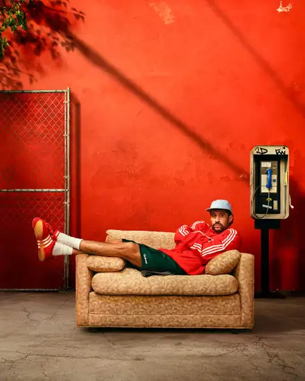 Editorial image of a person reclining on a vintage sofa outdoors while wearing red adidas sneakers and relaxed sportswear set against a vivid red wall