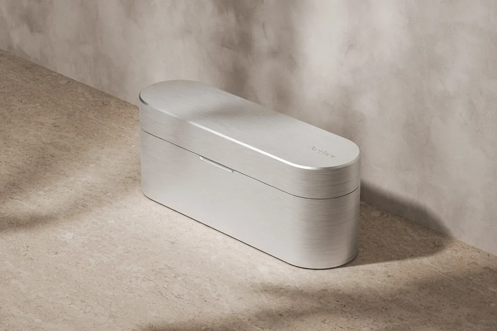A sleek, elongated metallic case with softly rounded edges sits on a textured stone surface, its brushed aluminum finish catching diffused light. The minimalist design is uninterrupted except for a subtle seam indicating the lid and a small engraved logo on top, giving it a refined, high-end, almost architectural presence against a neutral, softly backdropped