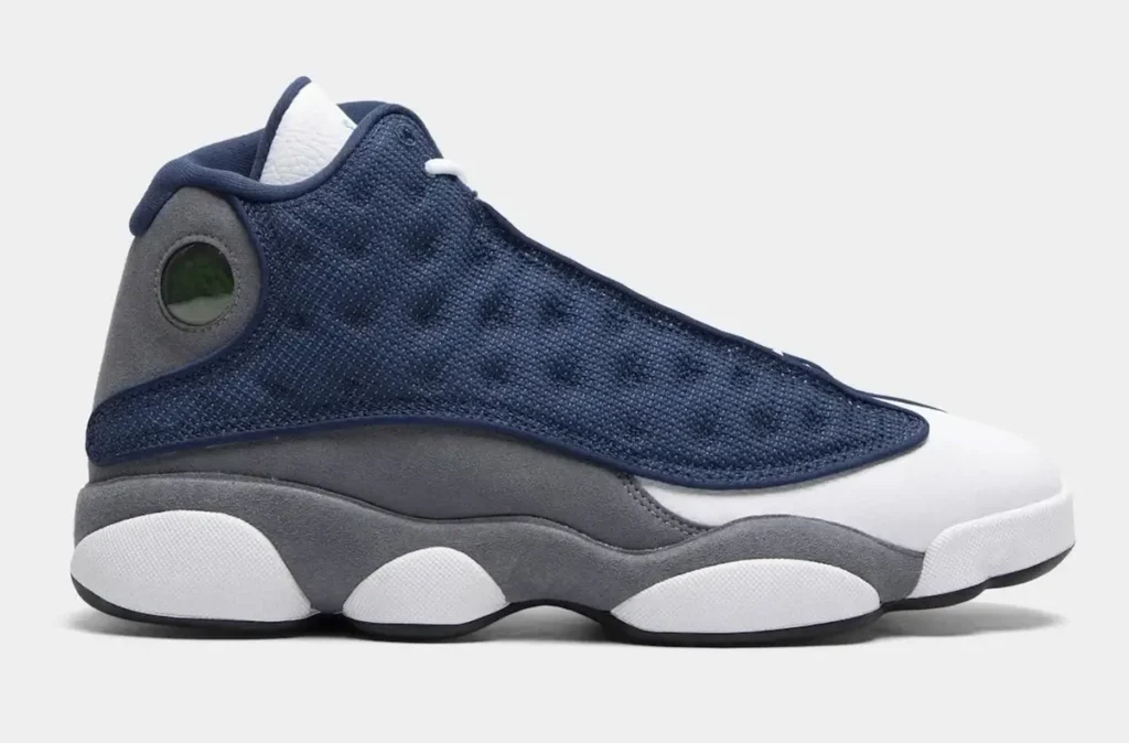 Side-profile view of the Air Jordan 13 in a navy, white, and grey color scheme featuring textured navy mesh paneling, white tumbled leather overlays, grey suede midsoles, and the signature green holographic heel emblem, displayed against a light grey studio background