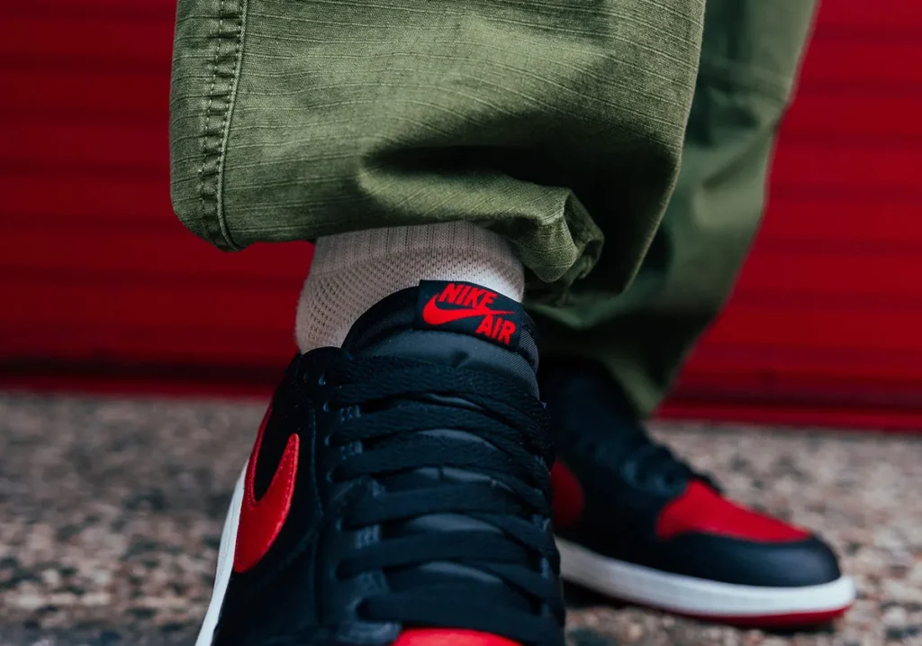 On-foot view of the Air Jordan 1 Low OG “Banned” showing black leather upper, varsity red Swoosh, and Nike Air tongue branding with olive pants styling