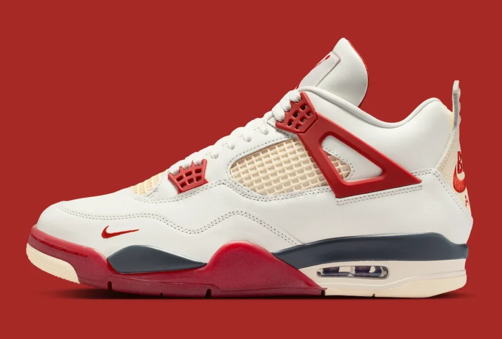 A lateral studio profile image of the Air Jordan 4 “Brick by Brick” displayed against a saturated red background. The shoe combines an off-white leather upper with cream mesh netting and bold red TPU support wings that define the iconic Jordan 4 framework. Matching red lace eyelets and forefoot detailing amplify the contrast, while a small embroidered red Swoosh near the toe introduces an additional heritage-inspired accent. Dark navy overlays wrap through the sculpted midsole above a visible Air cushioning unit, creating depth against the vintage cream outsole. At the heel, textured cream Nike Air branding reinforces the retro aesthetic, while the overall composition highlights the silhouette’s layered construction, premium material mix, and architectural “Brick by Brick” design language