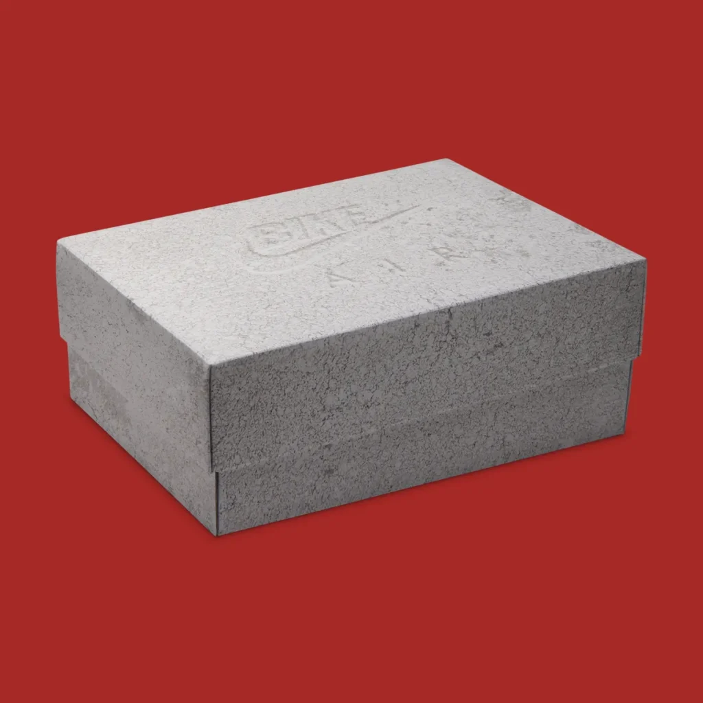 A macro-detail studio image highlighting the interior tongue construction of the Air Jordan 4 “Brick by Brick” against a rich red backdrop. The underside of the tongue is covered in plush red terry-style fabric, creating a textured contrast against the smooth off-white leather collar panels surrounding it. A stitched red fabric label appears at the center with inverted gray “JORDAN BIKING CO. 2017” lettering, referencing hidden archival-style branding details commonly associated with premium Jordan releases. Beneath the tongue, the cream heel tab panel features a heavily textured, almost weathered finish with raised circular embossing that reinforces the sneaker’s vintage-inspired material story and handcrafted aesthetic