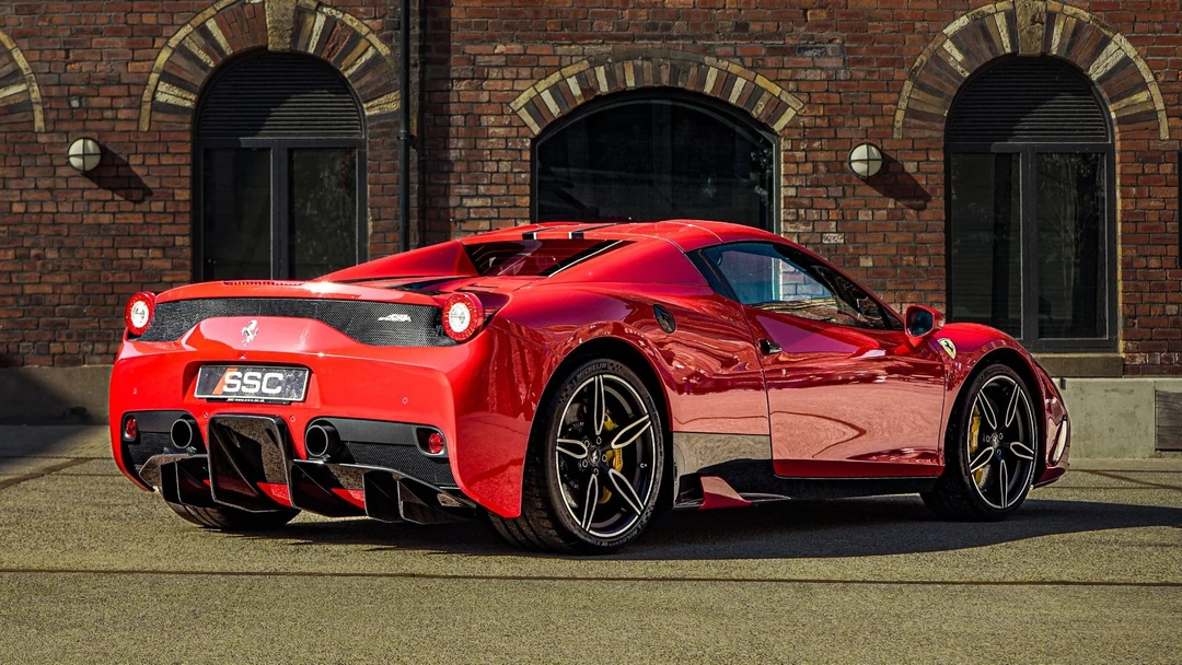 Rear three-quarter view of a red Ferrari 458 Italia parked on a street, showing sculpted bodywork, circular taillights, triple exhaust, and black diffuser against a brick building backdrop