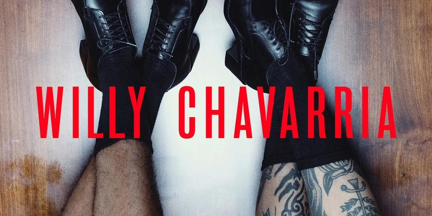 Close-up campaign-style image featuring two pairs of black leather lace-up boots positioned opposite each other on a wooden floor, with visible tattooed legs and bold red “WILLY CHAVARRIA” typography stretched across the center in a stark, cinematic fashion editorial composition
