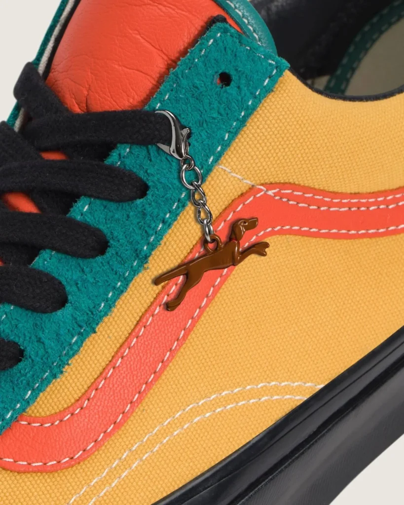 Close-up detail of an OTW by Vans shoe featuring mustard canvas panels, teal hairy suede overlays, coral leather side stripe accents, black laces, and a dangling brown dog-shaped metallic charm attached near the eyelets, highlighting the shoe’s textured premium craftsmanship
