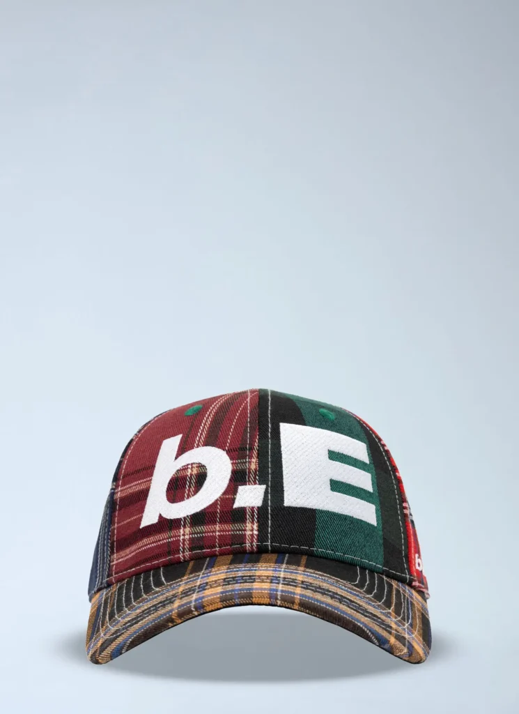 Front-facing studio product image of a patchwork logo baseball cap against a soft light grey background. The cap features mixed tartan and plaid fabric panels in burgundy, forest green, black, red, and navy, assembled into a quilt-like composition. Oversized white embroidered lettering spans the crown, creating a bold graphic contrast across the textured woven surfaces. The curved brim combines layered striped and plaid textiles, emphasizing handcrafted construction and a contemporary upcycled aesthetic