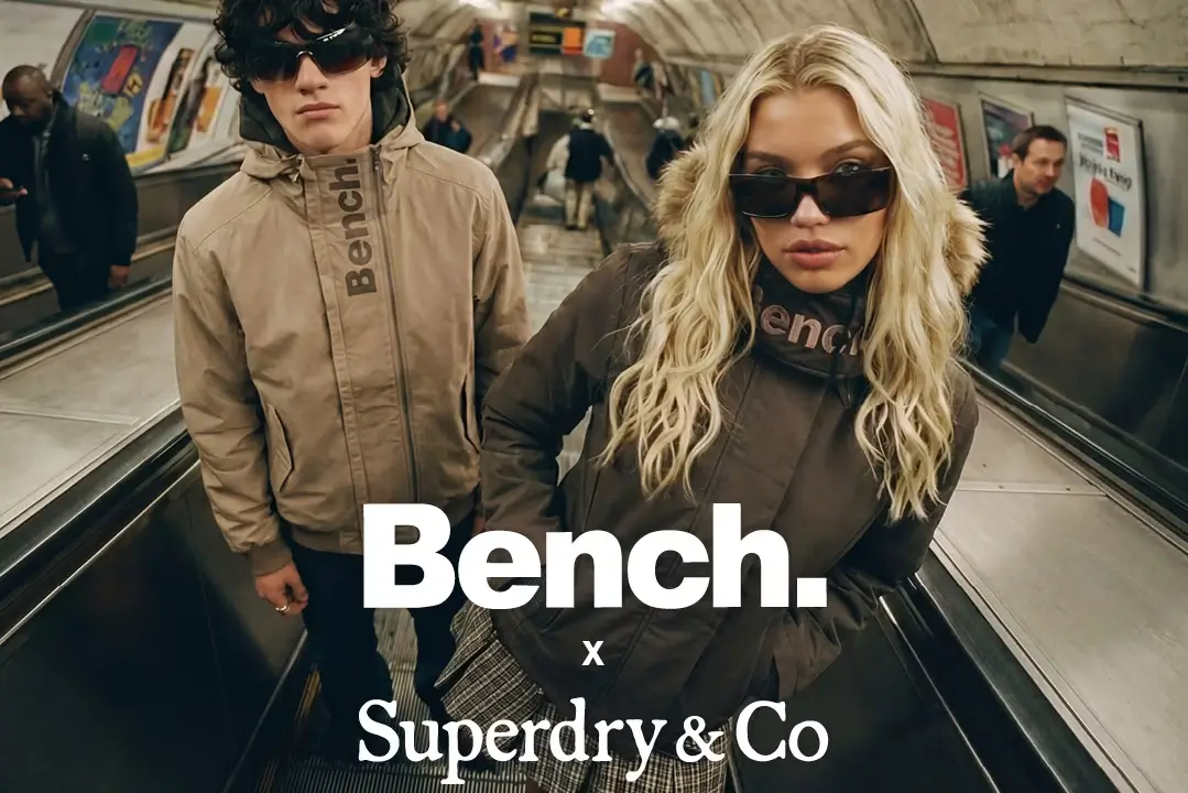 Two models wearing Bench outerwear stand inside a London Underground escalator tunnel beneath fluorescent lighting, capturing the gritty urban atmosphere of the Bench x Superdry & Co. campaign. One model wears a beige funnel-neck jacket with bold vertical Bench branding and oversized black sunglasses, while the other wears a dark fur-trimmed jacket with sleek sunglasses and platinum-blonde hair, reinforcing the collection’s refined Y2K streetwear aesthetic