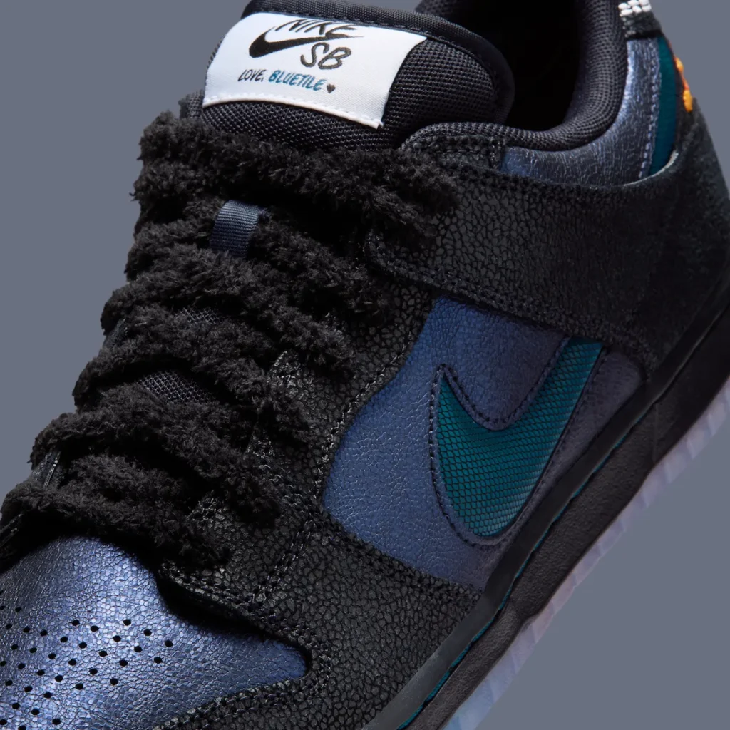 Close-up detail of the Bluetile x Nike SB Dunk Low “Monarch Butterfly” highlighting shaggy black fuzzy laces, cracked-texture black overlays, shimmering navy leather underlays, and a textured Marina blue Swoosh with custom Nike SB Bluetile tongue branding